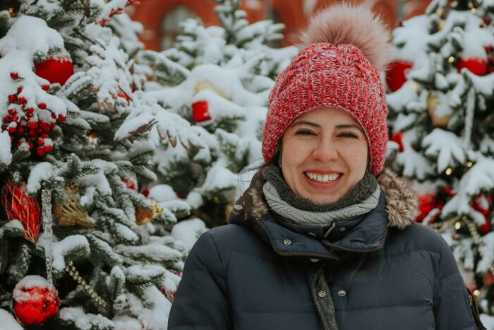A person smiling in front of snow-covered Christmas trees, wearing a red knit hat and a dark winter coat. Festive and joyful winter scene.