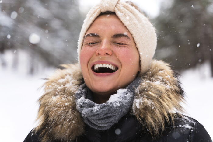 A woman joyfully laughs in a snowy landscape, wearing a furry hooded coat, scarf, and headband. Snowflakes gently fall around her, enhancing a cheerful mood.